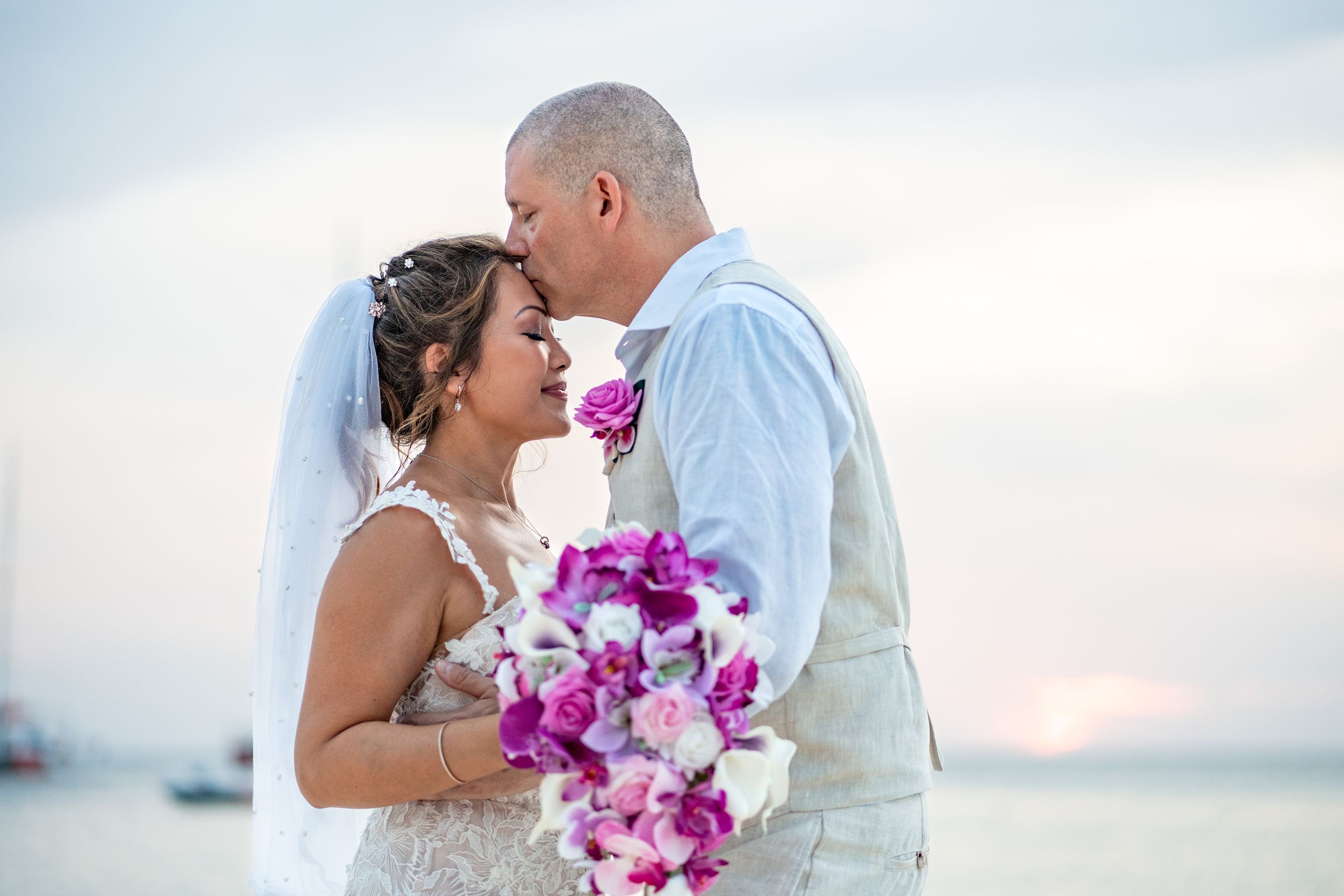Groom kissing bride on forehead while bride hold vibrant purple bouquet.